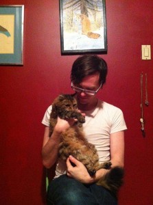Clint Enns sits in front of a red wall while holding his fluffy cat Mia