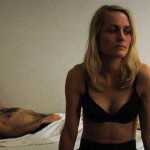 Man lies in bed while woman in bra looks forlorn