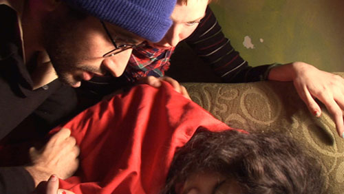 Man wearing winter hat tries to wake up a sleeping woman