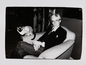 Artist Andy Warhol sits in a chair with a siamese cat