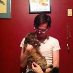 Clint Enns sits in front of a red wall while holding his fluffy cat Mia