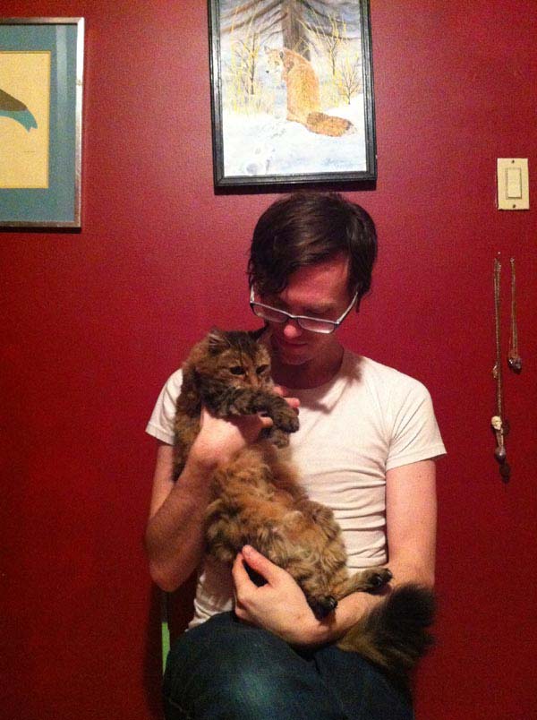 Clint Enns sits in front of a red wall while holding his fluffy cat Mia