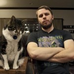 Filmmaker Brady Hall, wearing a Megadeth T-shirt, sits with his arms crossed next to his large cat Reggie
