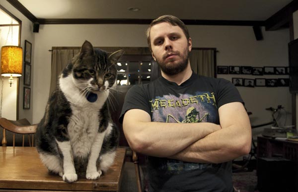 Filmmaker Brady Hall, wearing a Megadeth T-shirt, sits with his arms crossed next to his large cat Reggie