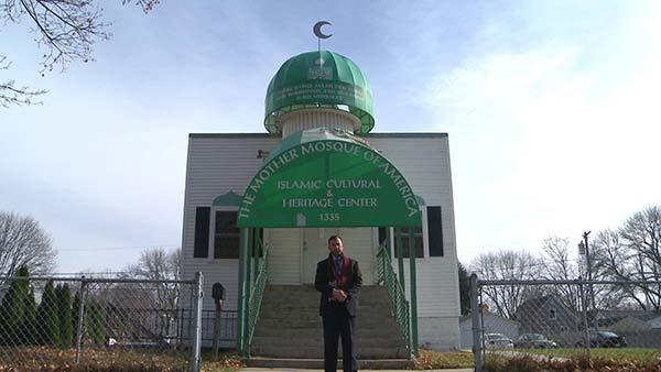 An imam stands in front of his mosque with a green awning