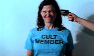 Woman wearing a t-shirt that says "Cult Member" has a gun pointed at her head