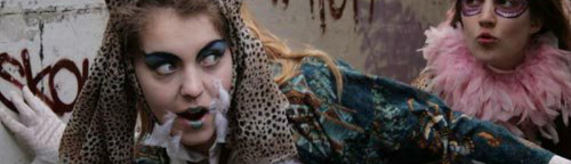 Two teenage girls dressed in outlandish costumes run down a dirty alley